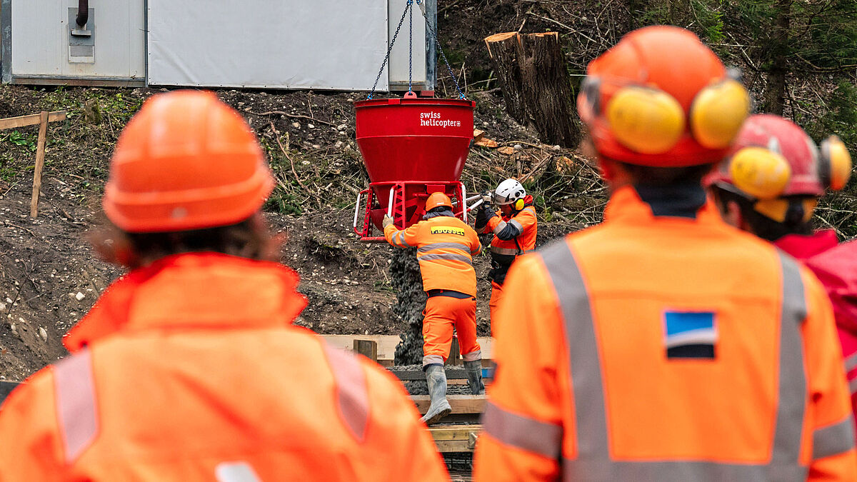 La classe de maçons, aidée par des apprentis d’autres professions, en train de vider le béton pour les fondations. La benne à béton est suspendue à l’hélicoptère.